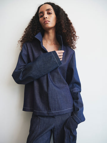 A person with curly hair poses against a white background, wearing Zero + Maria Cornejo's Denim Biker Shirt. Made from organic denim and GOTS-certified cotton, this stylish piece with a wide collar and long sleeves pairs perfectly with cropped pants for a casual yet chic look.