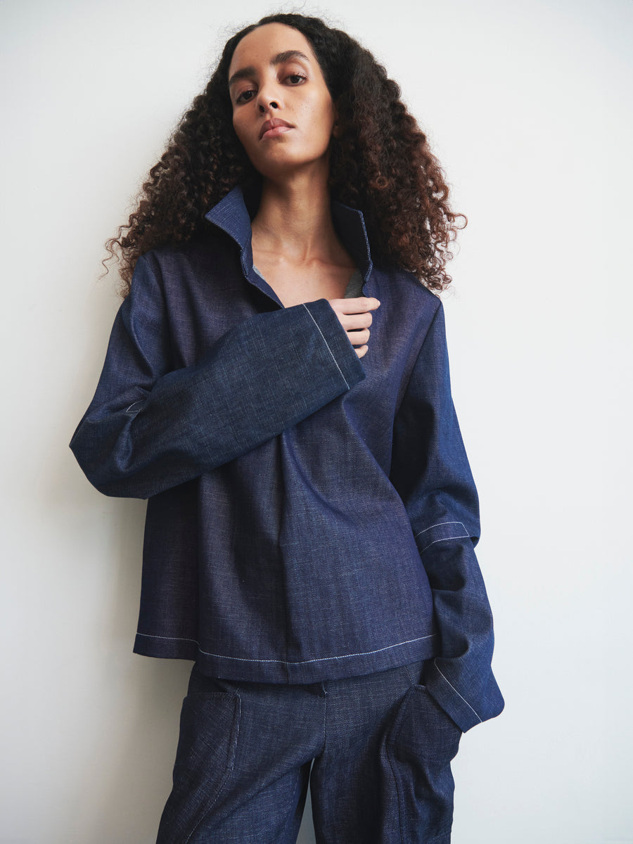 A person with curly hair poses against a white background, wearing Zero + Maria Cornejo's Denim Biker Shirt. Made from organic denim and GOTS-certified cotton, this stylish piece with a wide collar and long sleeves pairs perfectly with cropped pants for a casual yet chic look.