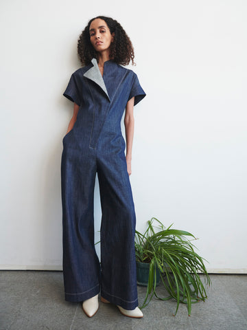 A person with long, curly hair poses against a white wall, wearing the Edi Boiler Suit by Zero + Maria Cornejo—a GOTS-certified organic denim outfit—and stylish white pointed shoes. A green potted plant adds a touch of nature to the gray floor in the background.