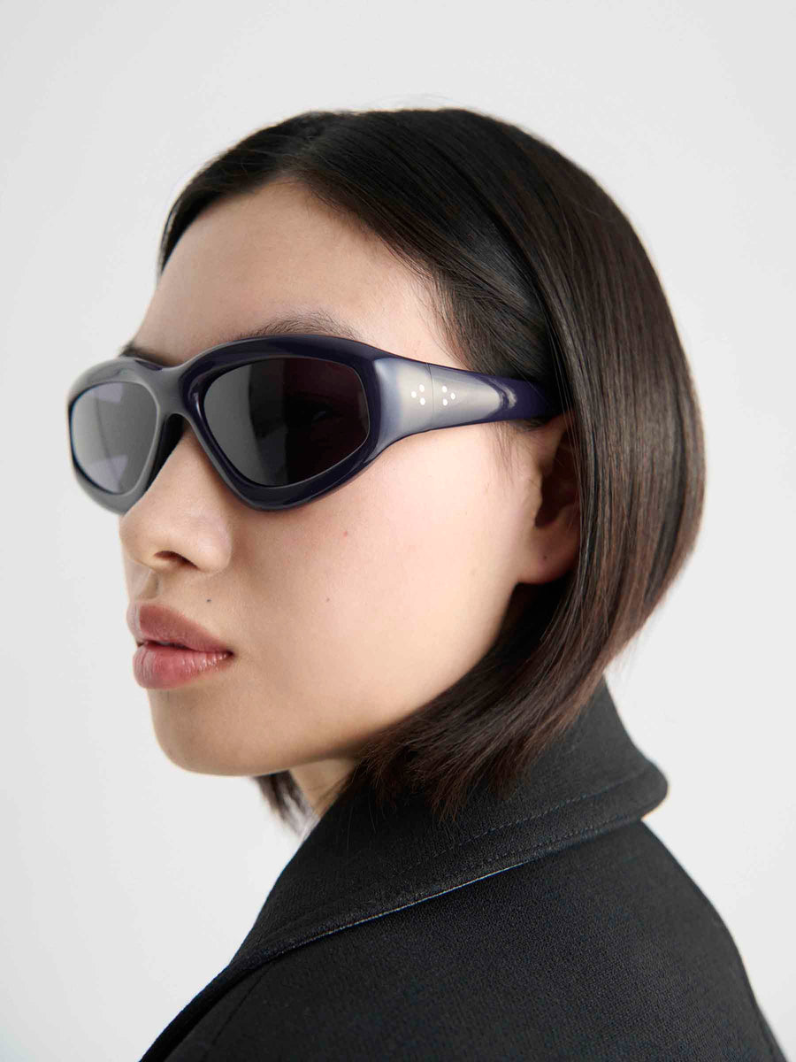 A person with straight, dark hair and a bob wears Port Tanger Irfan unisex eyewear by Port Tanger, featuring UVA/UVB protection and a black jacket, looking over their shoulder against a plain white background.