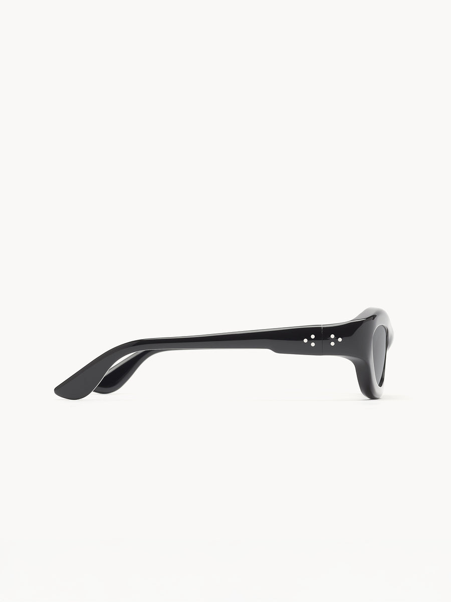 The Port Tanger Hayya by Port Tanger are black cat-eye sunglasses with a thick frame and temples, featuring two small circular metallic accents near the hinge. Set against a white background, they offer classic style with UVA/UVB protection.