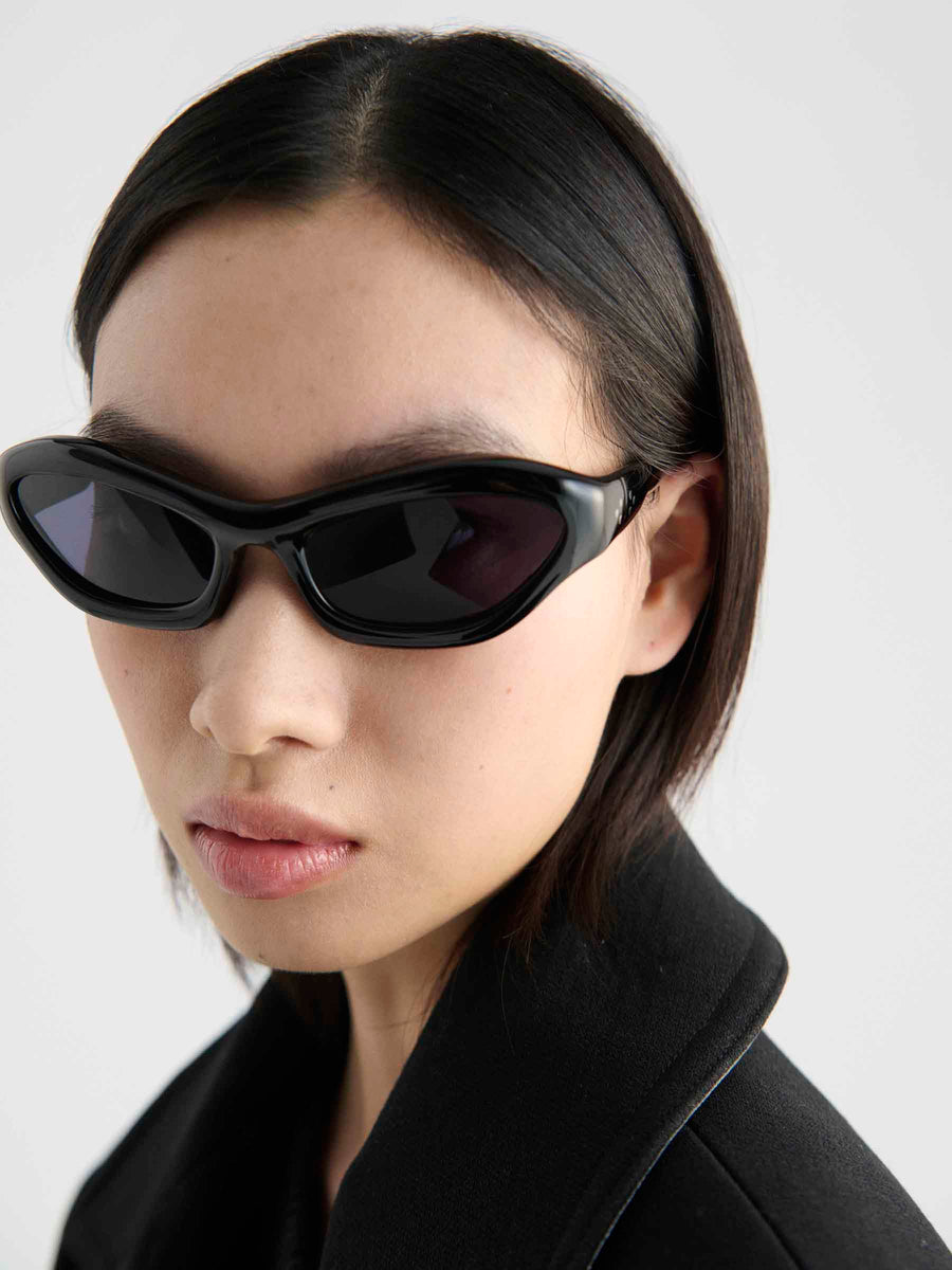 A person with straight, chin-length dark hair wears black Port Tanger Hayya unisex eyewear from Port Tanger with UVA/UVB protection and a black jacket, looking slightly to the side against a plain light background.
