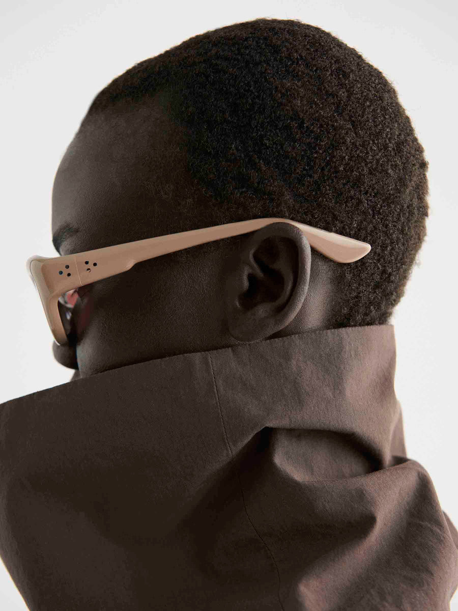 A person with short curly hair wears Port Tanger Hayya unisex eyewear with light-colored frames and a high-collared brown jacket, shown in profile against a plain background.