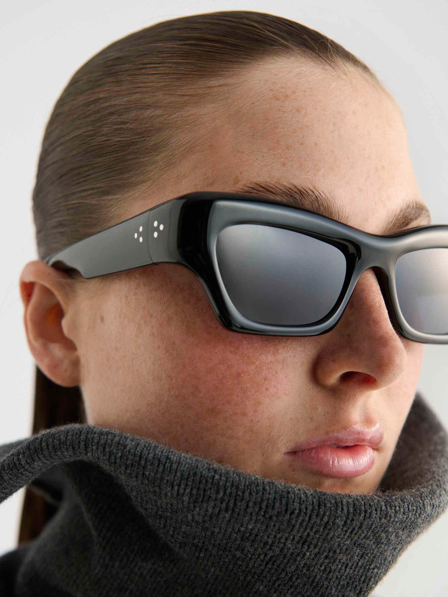 A person with slicked-back hair wears Port Tanger Ayreen oversized black sunglasses by Port Tanger, handcrafted in Japan with small white dots on the frames, and a high-collared dark grey sweater, looking forward with a neutral expression.