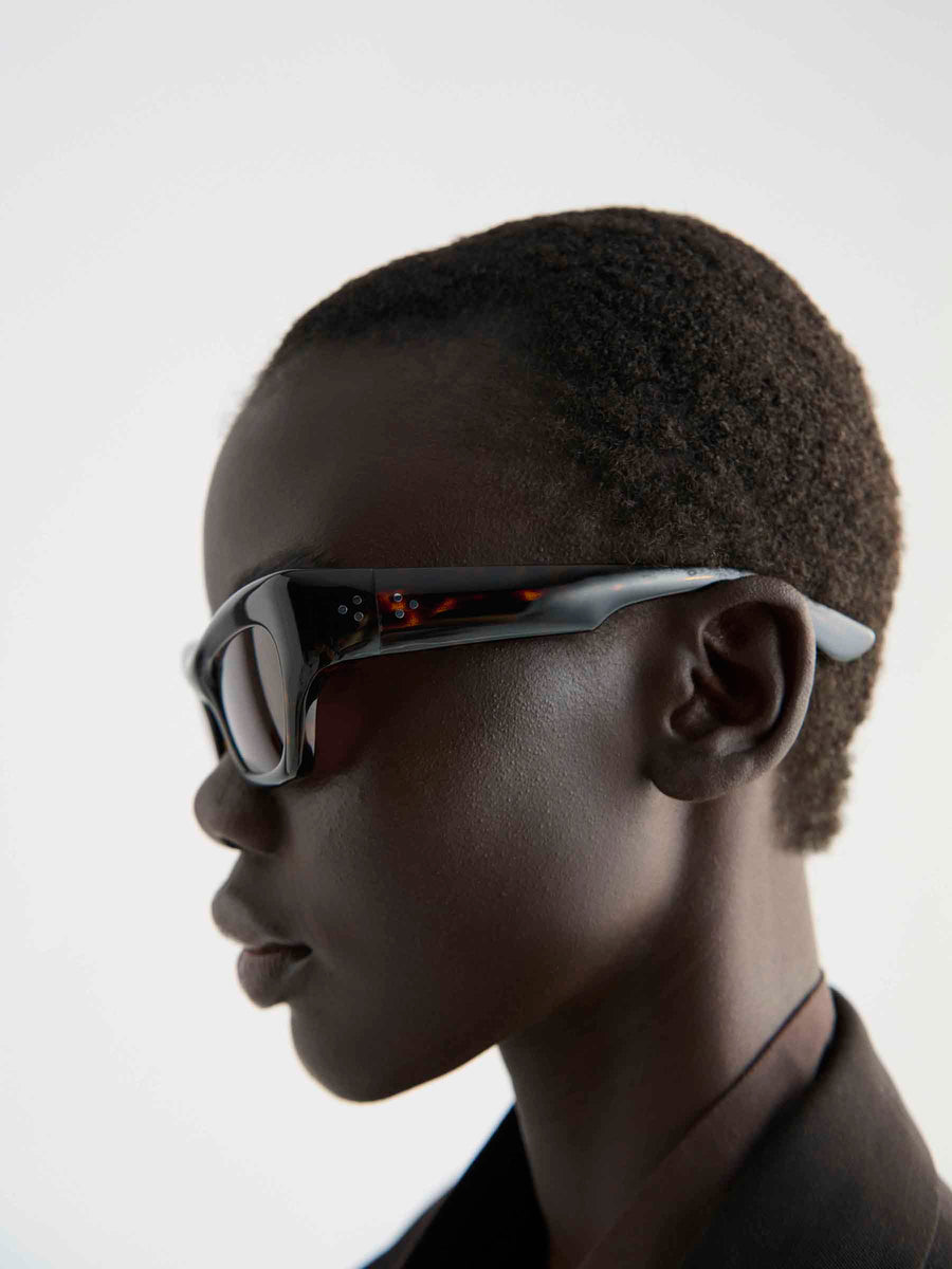 Profile view of a person with short curly hair wearing Port Tanger Ayreen cat-eye sunglasses by Port Tanger, featuring thick dark frames and 100% UVA/UVB protection, paired with a dark jacket against a plain light background.