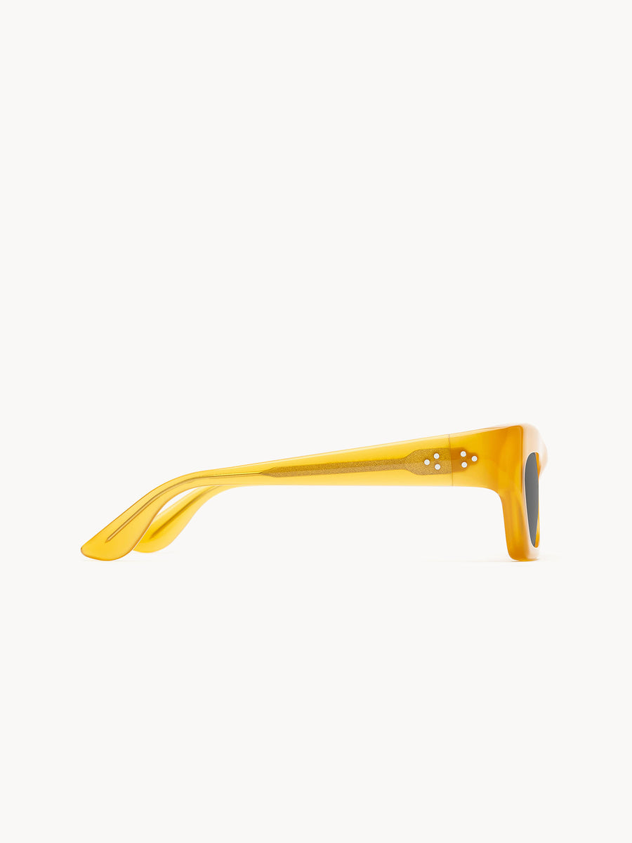 Side view of Port Tanger Ayreen chunky, rectangular yellow-orange sunglasses by Port Tanger, featuring dark lenses, three small white dots near the hinge, and 100% UVA/UVB protection on a plain white background.