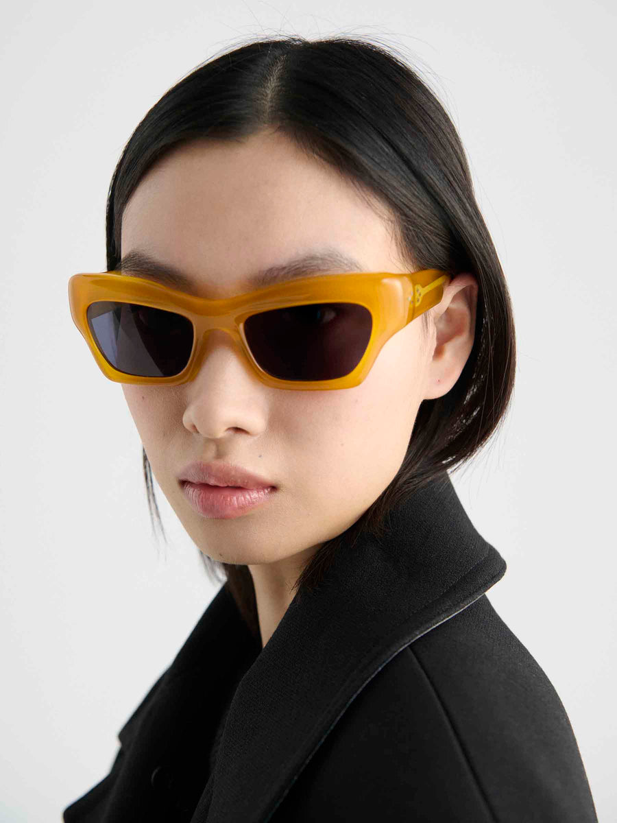 A person with straight black hair wears Port Tanger Ayreen yellow rectangular sunglasses by Port Tanger and a black coat, looking confidently at the camera against a plain light background.