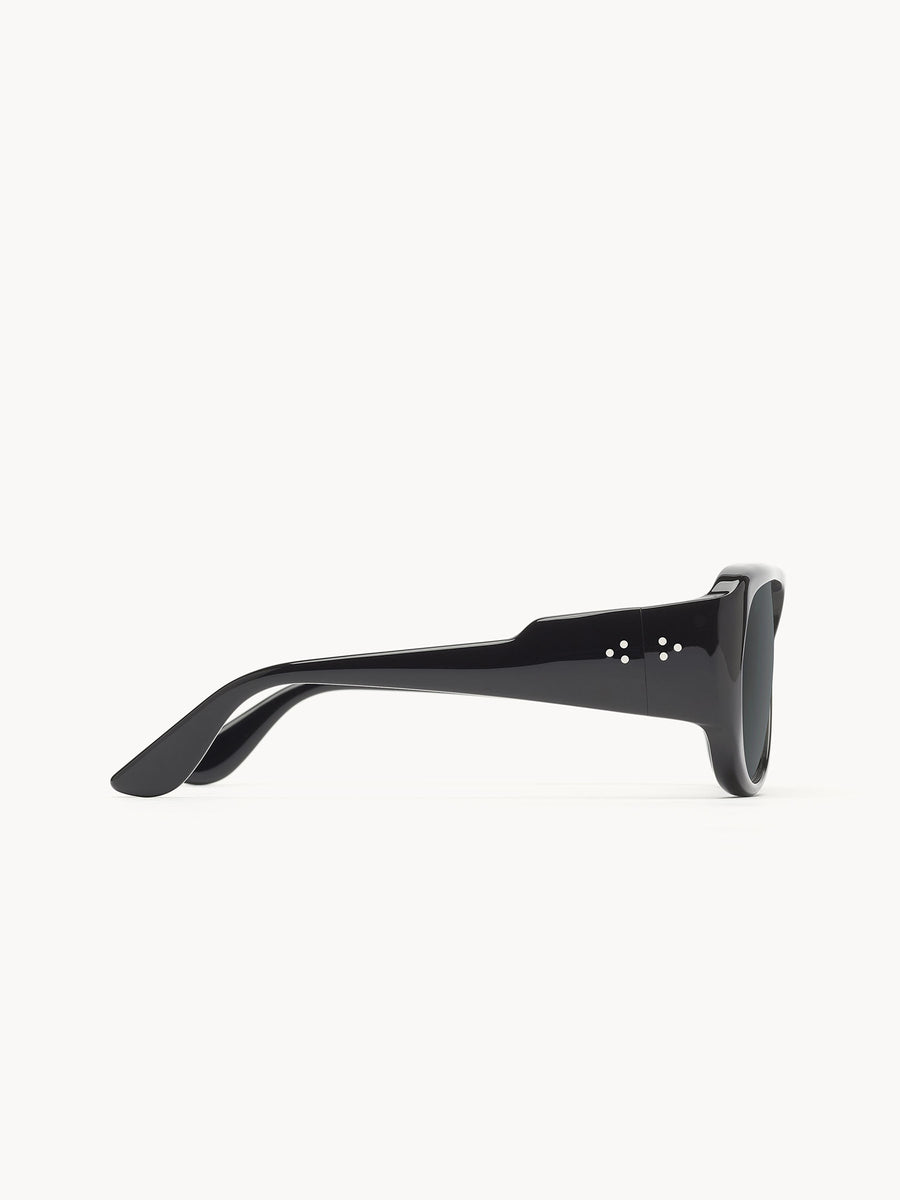 Side view of Port Tanger Gambia black sunglasses by Port Tanger, featuring thick glossy frames, dark lenses with UVA/UVB protection, and two small circular accents on the arms. Handcrafted in Japan, shown on a white background.