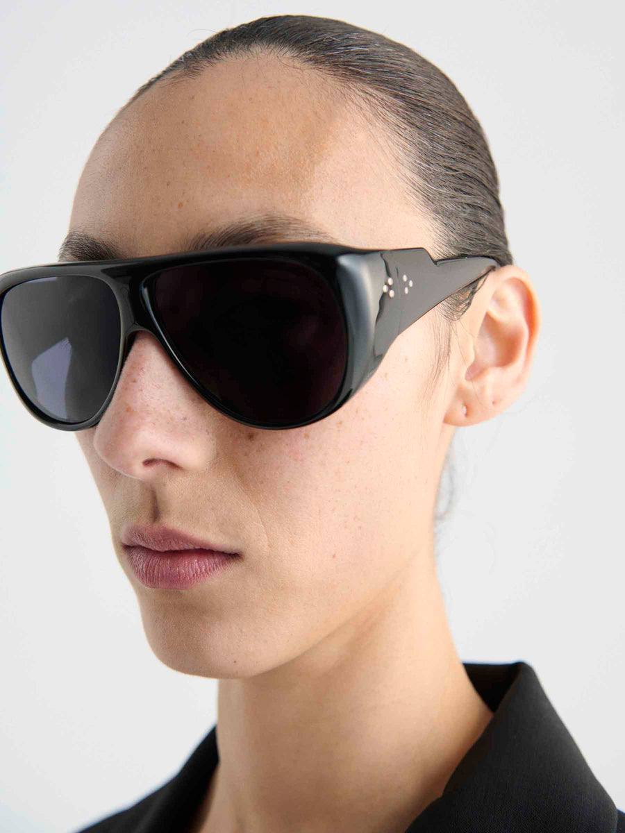 A person with slicked-back dark hair wears Port Tanger Gambia sunglasses by Port Tanger, featuring large black aviator frames and a black collar. The background is plain and light-colored, and the person's expression is neutral.