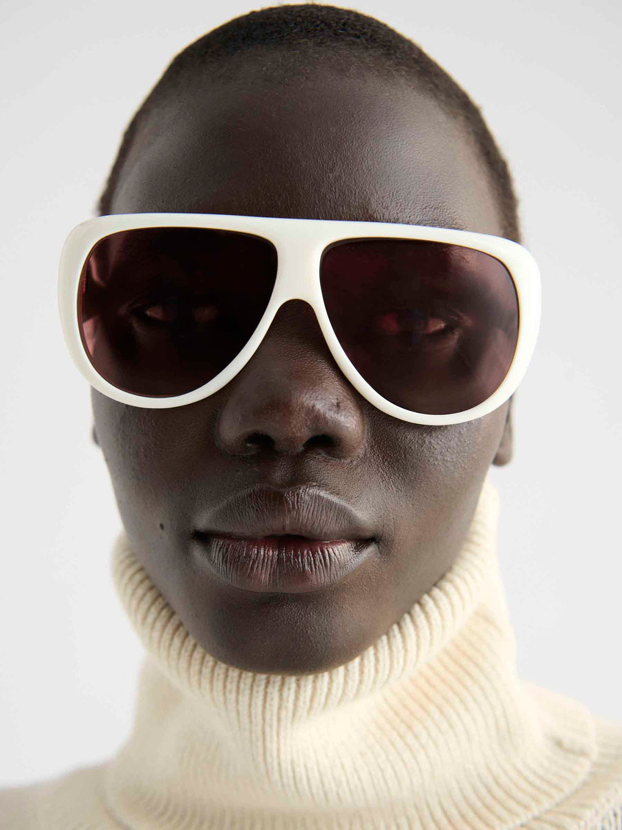 A person with dark skin wears Port Tanger Gambia sunglasses by Port Tanger, which feature large white frames and UVA/UVB protection, along with a cream ribbed turtleneck sweater, looking directly at the camera with a neutral expression.