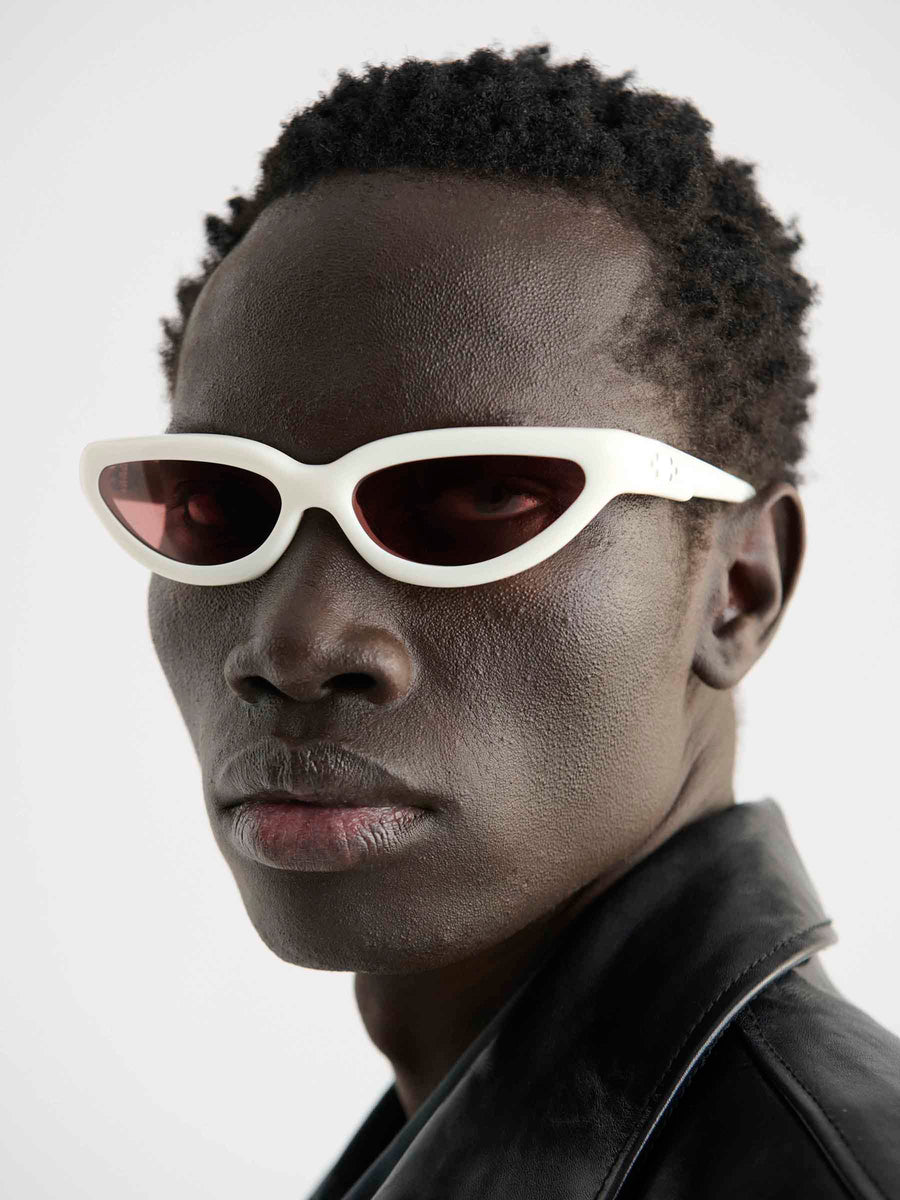 A person with short curly hair wears Port Tanger Fanna by Port Tanger—white cat-eye sunglasses with 100% UVA/UVB protection—paired with a black leather jacket, looking straight at the camera against a plain backdrop.