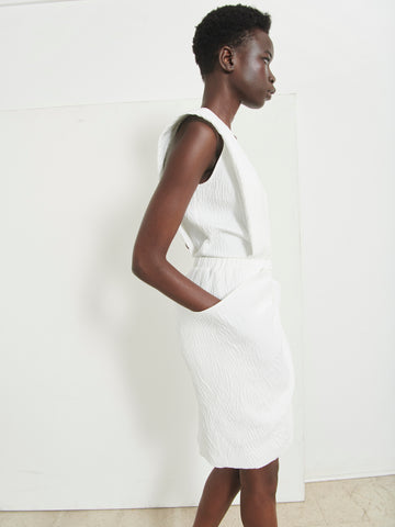 A person stands in profile against a plain white background, wearing a sleeveless, quilted above-knee Mackie Dress with pockets by Zero + Maria Cornejo. The dress has a fitted waist and knee-length hem, and the individual has short, curly black hair.