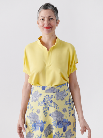 A person with short gray hair smiles against a light background, wearing a yellow draped sleeve Mare Shirt - Final Sale and a skirt with blue and gray floral patterns, both New York made by Zero + Maria Cornejo.