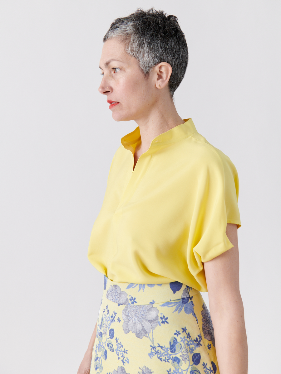 A short gray-haired individual stands sideways against a white background, wearing the Mare Shirt by Zero + Maria Cornejo. The shirt is bright yellow with draped sleeves, paired with a matching yellow skirt featuring blue floral patterns.