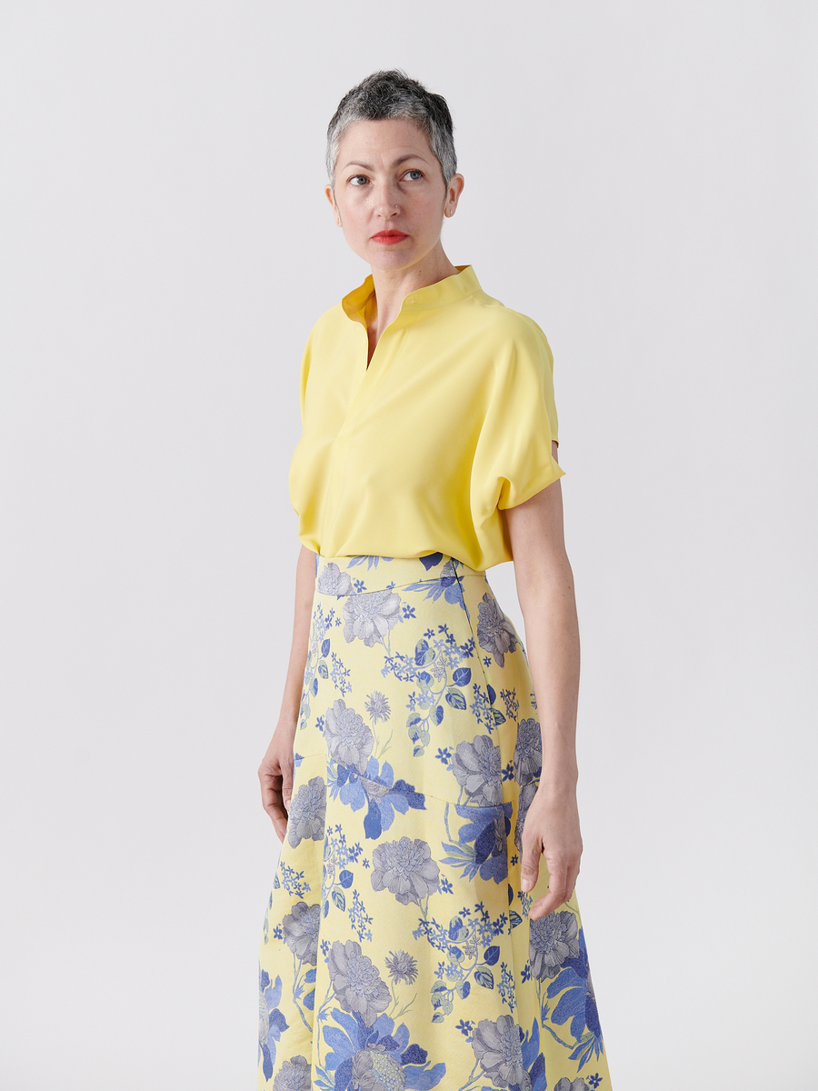 A woman stands against a plain backdrop in the Mare Shirt from Zero + Maria Cornejo, featuring draped sleeve details, and a matching skirt with blue floral patterns. Her short grey hair and neutral expression embody the elegance of New York-made design.