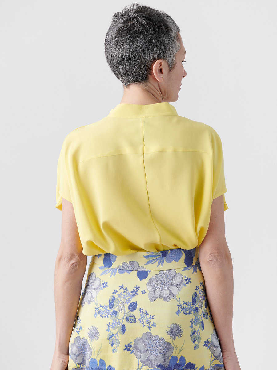 A person with short gray hair faces away from the camera, showcasing the New York-made Mare Shirt by Zero + Maria Cornejo, which features draped sleeves. They pair it elegantly with a yellow skirt adorned with a blue floral pattern against a minimalist white background.