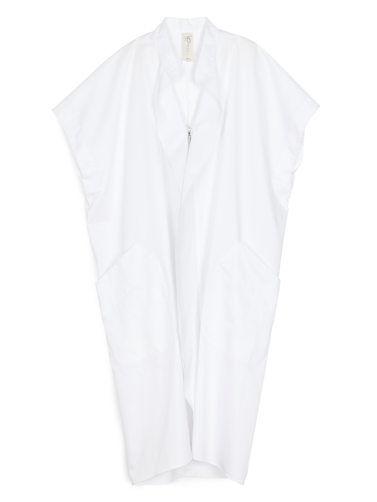 In a minimalist room, a person with curly hair stands confidently wearing the Fin Gaban Dress by Zero + Maria Cornejo, featuring subtle ruffle detail and paired with black slip-on shoes. A dress form is visible in the background.