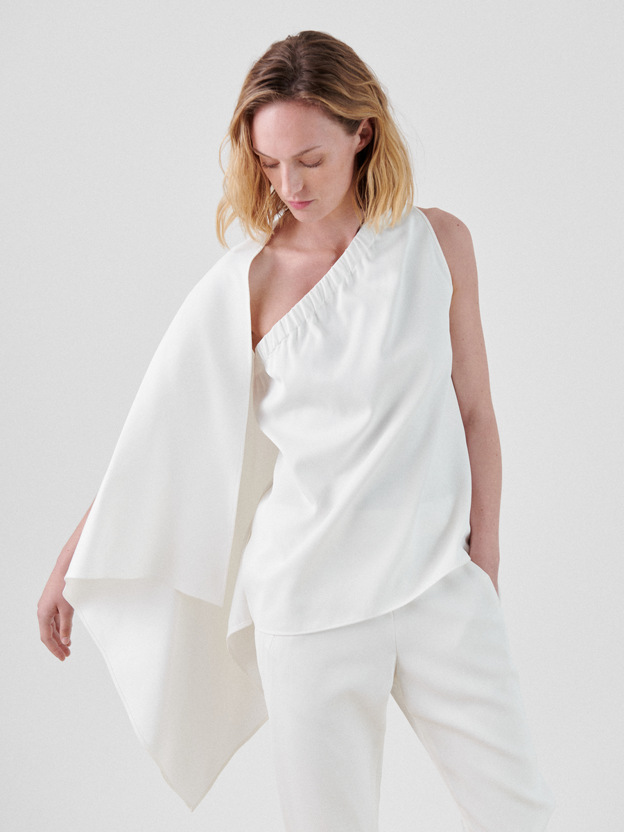 A woman with blonde hair is wearing an elegant white asymmetrical neckline one-shoulder One Top by Zero + Maria Cornejo, made from FSC certified viscose, featuring a flowing drape on one side, paired with white pants. She is looking down with one arm relaxed by her side and the other arm bent slightly at the elbow. The background is plain white.