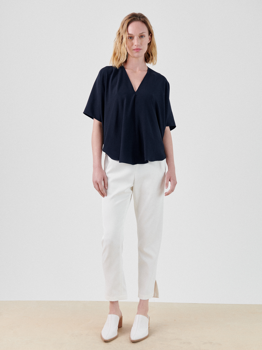 A person with shoulder-length blond hair wearing a loose-fitting, short-sleeve black Circle Top by Zero + Maria Cornejo paired with white pants and white shoes stands against a plain white background. The person's expression is neutral, and they are looking directly at the camera.