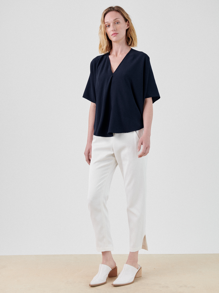 A person with light hair stands against a plain background. They are wearing a navy blue V-neck top with short sleeves, Slim Elli Pant by Zero + Maria Cornejo featuring hidden pockets and an elastic waist, and white slip-on shoes with a low heel. The person is looking forward with a relaxed posture.