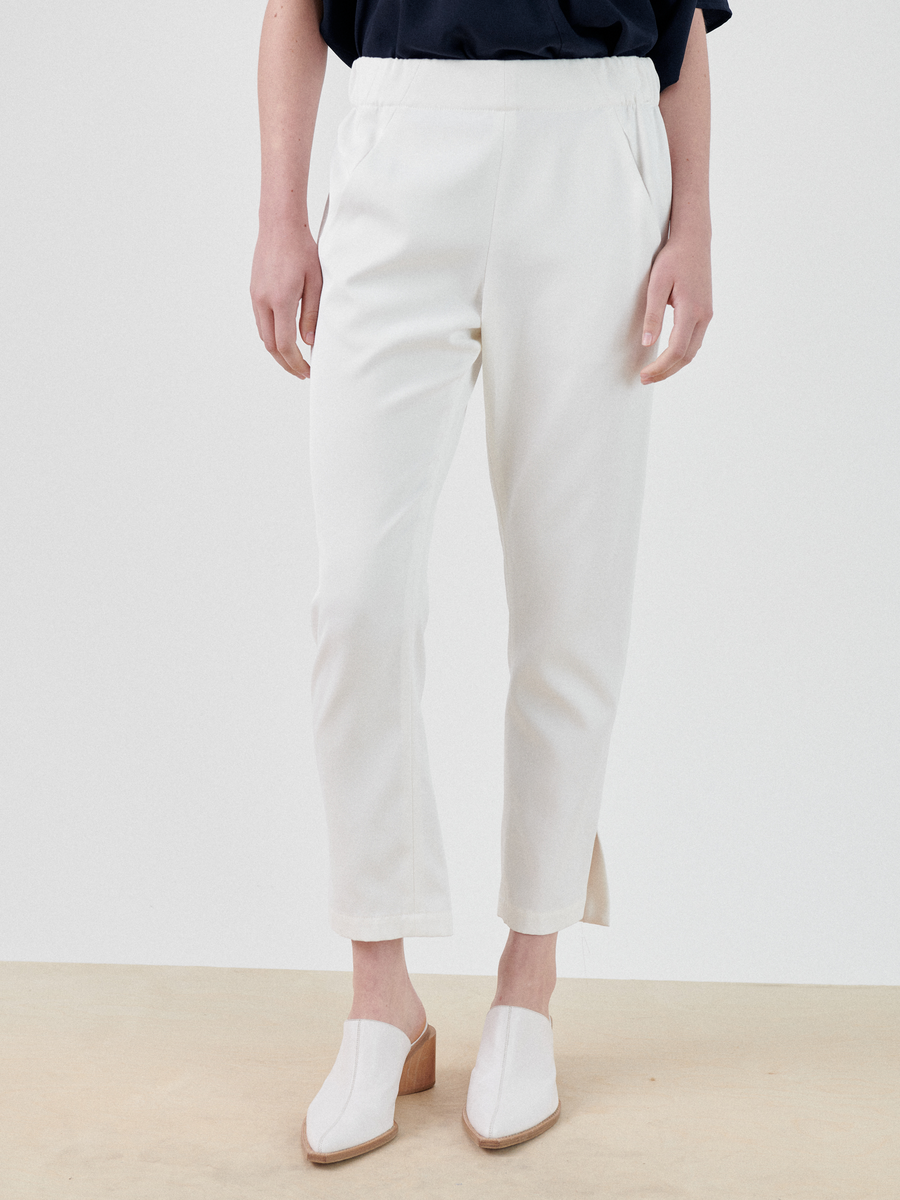 A person is standing wearing white Slim Elli Pants by Zero + Maria Cornejo with side slits near the ankles and white mule shoes with a wooden heel. The top of their outfit is a loose-fitting dark short-sleeve shirt. The background is plain and neutral.