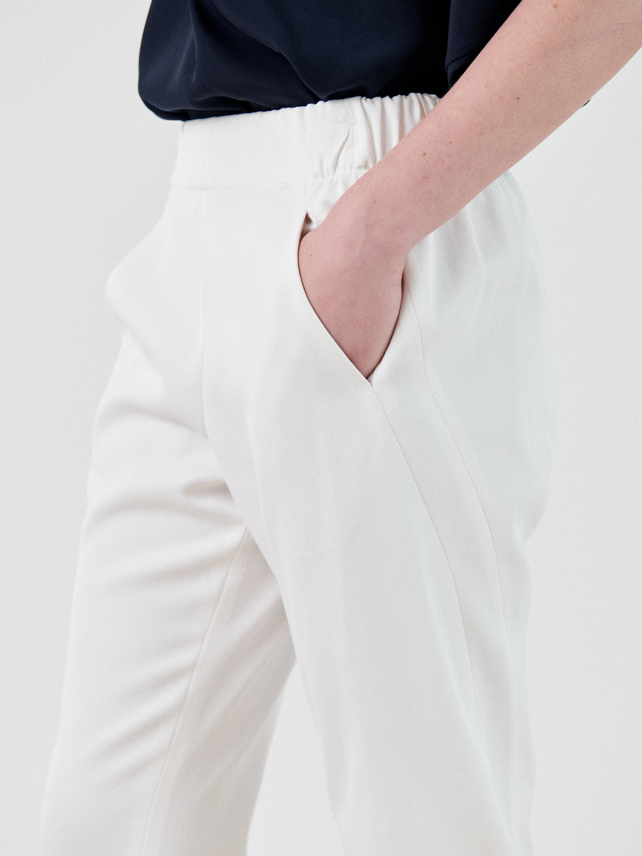 Close-up of a person wearing white Slim Elli Pant by Zero + Maria Cornejo with an elastic waistband and hidden side pockets, with one hand in the pocket. The person is also wearing a dark top. The image focuses on the lower torso and part of the upper thigh, without showing the full body.