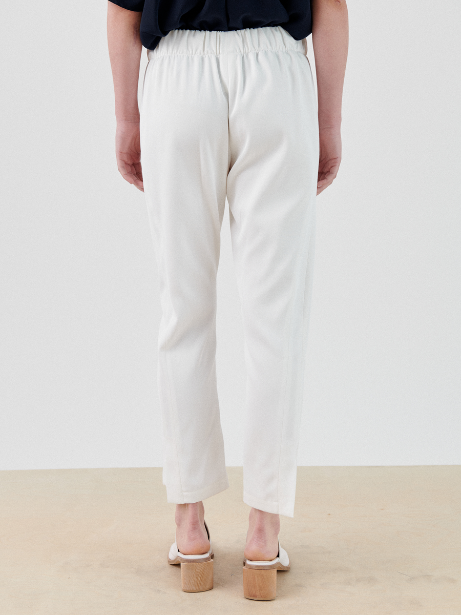 A person is standing with their back facing the camera, wearing white, high-waisted Slim Elli Pant from Zero + Maria Cornejo featuring an elastic waist and hidden pockets. They are also wearing a dark top and light-colored heeled sandals. The background is plain and light-colored.