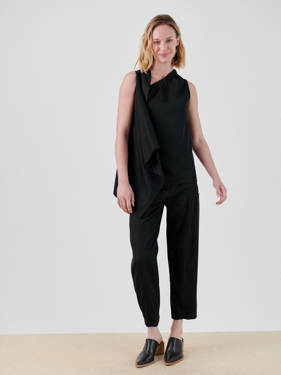 A woman with shoulder-length blonde hair stands smiling against a plain background. She is wearing a sleeveless black top that drapes over one shoulder, the Margot Pant by Zero + Maria Cornejo, and black slip-on shoes made of viscose-cotton twill. She is positioned with one arm slightly bent and the other straight at her side.