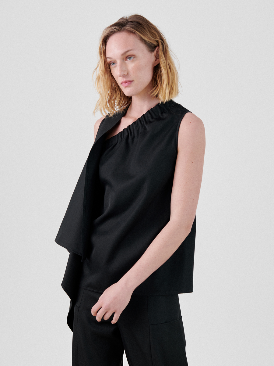 A person with shoulder-length blonde hair is wearing the One Top, a black asymmetrical sleeveless top with a ruffled neckline made from FSC certified yarns by Zero + Maria Cornejo. They stand against a plain gray background, gazing slightly to the left.