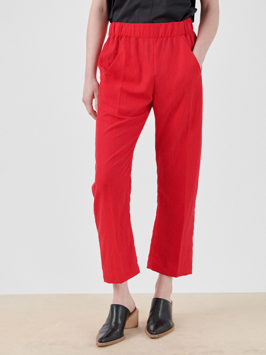 A person is standing, wearing bright red Zero + Maria Cornejo Eko Pant with an elastic waistband. Their hands are in the hidden pockets. They are also wearing a black top and black slip-on shoes with a tinge of brown at the bottom. The background is light gray.