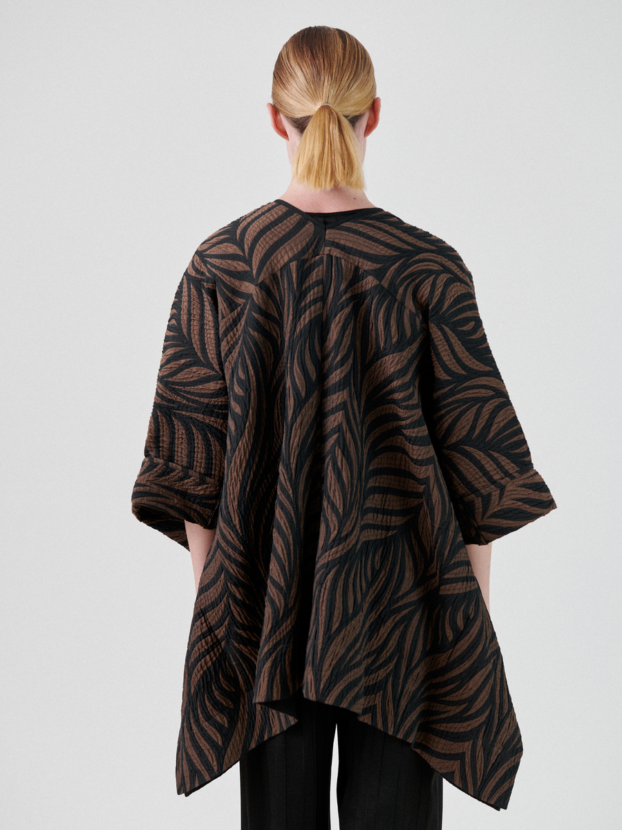 A person with light brown hair tied in a low ponytail is seen from behind, wearing a loose, long-sleeved top featuring a wavy black and brown leaf-like pattern. The Eve Square Shrug by Zero + Maria Cornejo, made from organic cotton, has a flowing asymmetrical hem and is paired with black pants.