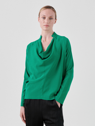 A person with short, light-colored hair is wearing a loose-fitting green silk charmeuse Zero + Maria Cornejo Long-Sleeved Evi Top with a draped neckline and black trousers. The background is plain white. The person is looking directly at the camera with a neutral expression.