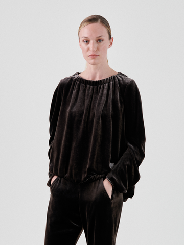 A person with a neutral expression wears the Ruched Bubble Top by Zero + Maria Cornejo, featuring a luxurious velvet fabric. The outfit's subtle iridescent sheen contrasts against the plain white background as they pose with hands in pockets and hair tied back neatly.