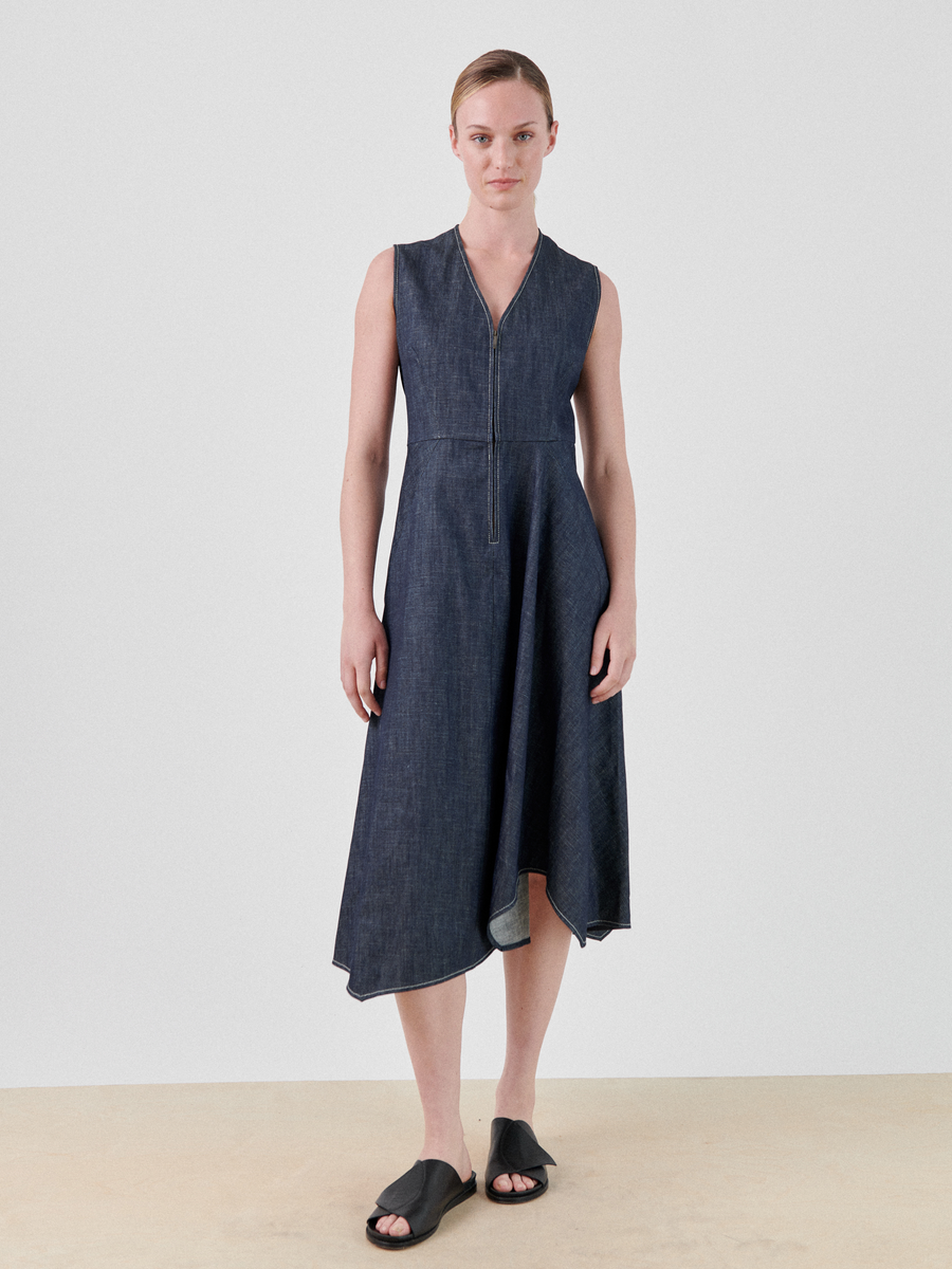 A person stands against a plain white background, wearing a sleeveless, dark denim dress with a V-neckline and zipper detail. The GOTS-certified organic **Denim Wave Dress** by **Zero + Maria Cornejo** has an asymmetrical hemline. They are also wearing black slide sandals and have their hair tied back.