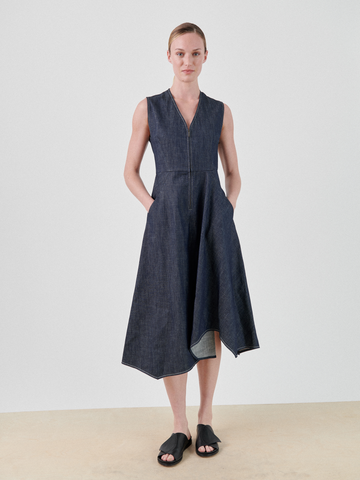 A person with their hair tied back is standing against a plain background, wearing a sleeveless, V-neck, dark Denim Wave Dress from Zero + Maria Cornejo with an asymmetrical hem. Made from 100% organic cotton and GOTS-certified, the dress features handy pockets for their relaxed hands. They are also wearing black open-toed sandals.