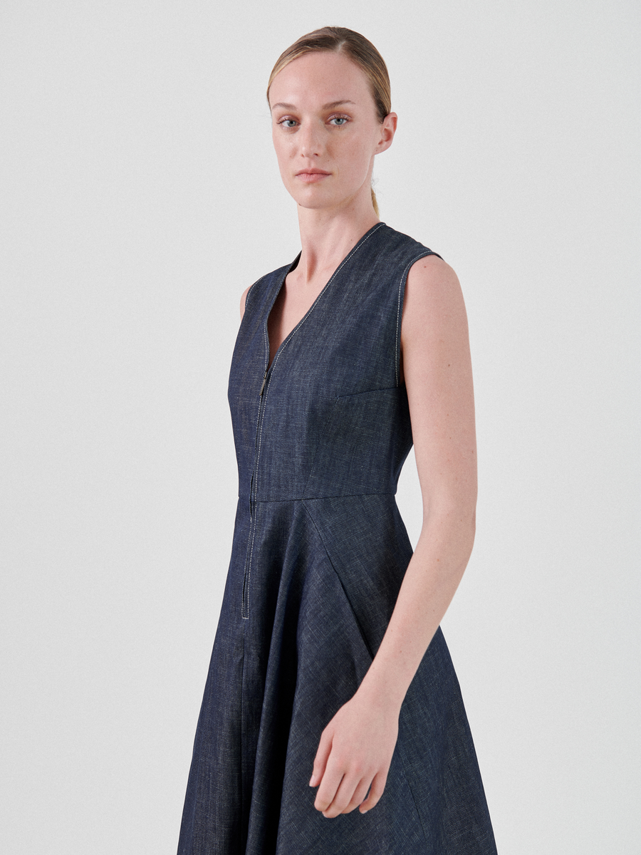 A person with light hair stands against a plain background wearing a Zero + Maria Cornejo Denim Wave Dress made from 100% organic cotton that has a V-neck and fitted bodice flaring into a wide skirt. They face slightly to the side with a neutral expression.