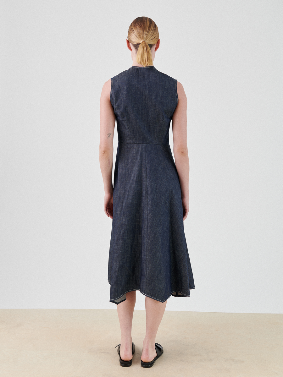 A person with long, blonde hair tied back in a ponytail is standing facing away from the camera. They are wearing a sleeveless, dark blue Denim Wave Dress by Zero + Maria Cornejo with a handkerchief hemline and black slip-on shoes. The background is a plain, light-colored wall and floor.