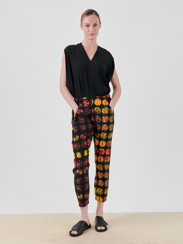 Against a plain backdrop, someone wears a black sleeveless top and Zero + Maria Cornejo's Gabi Trouser - Final Sale, featuring red, yellow, and orange circles. Their sustainable screen-printed outfit is completed with black slide sandals as they casually rest their hands in pockets.