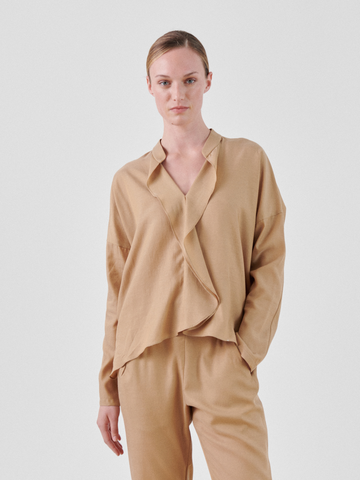 A person with light skin and short, pulled-back hair is wearing a beige Long-Sleeved Fin Gaban Shirt by Zero + Maria Cornejo and matching pants. They have one hand in their pocket and are standing against a plain light gray background, looking directly at the camera with a neutral expression.