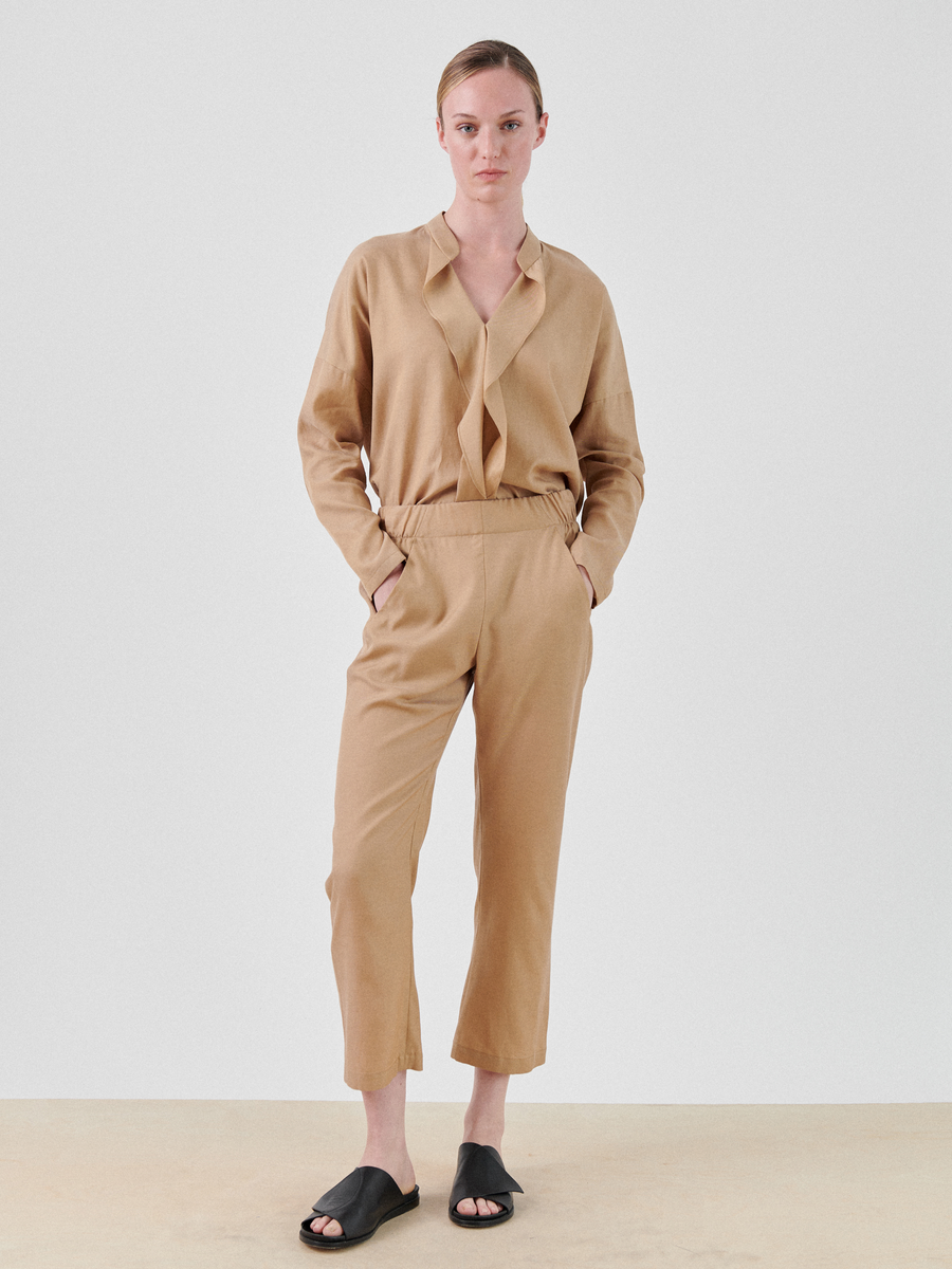 A person is standing against a plain white background. They are wearing a tan, loose-fitting outfit crafted from an eco-friendly cupro cotton blend with a long-sleeve top and soft draped Eko Pant by Zero + Maria Cornejo. Their hands are in their pockets, and they are wearing black slide sandals. The expression is neutral.