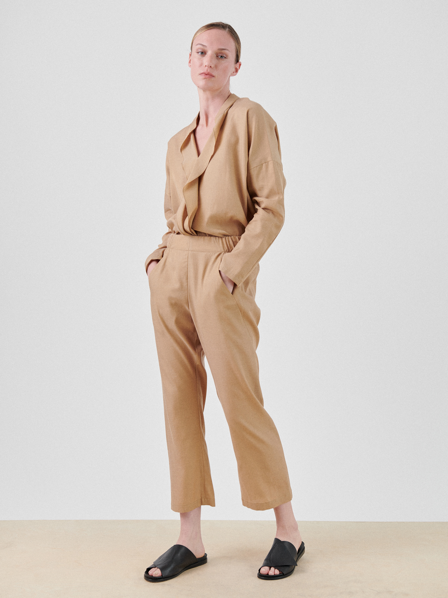 A person with short hair is standing against a plain, white background, dressed in a beige, loose-fitting **Eko Pant** from **Zero + Maria Cornejo** made from a cupro cotton blend with hands in pockets. They are wearing black, open-toe slide sandals and have a neutral expression on their face.