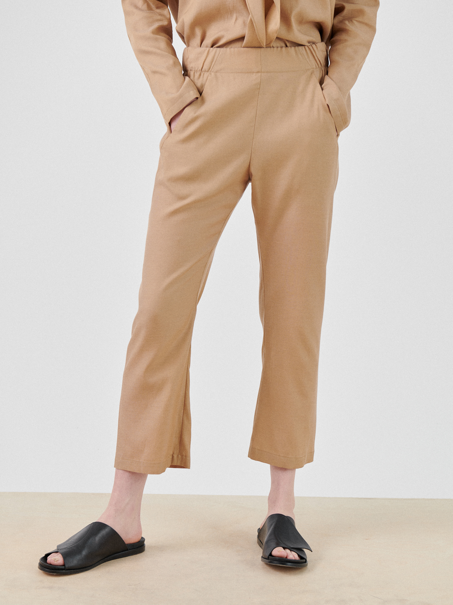 A person wearing tan ankle-length Eko Pant made by Zero + Maria Cornejo, made from a cupro cotton blend and a matching long-sleeve top. The person is standing with hands in pockets, and is wearing black open-toe slide sandals. The background is neutral.