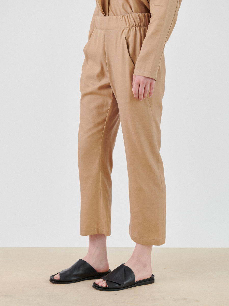 A person wearing tan, loose-fitting, soft draped Zero + Maria Cornejo Eko Pant and a matching long-sleeve top stands against a plain background. They are also wearing black slide sandals with an open-toe design. The image is cropped, showing only the lower half of the body.