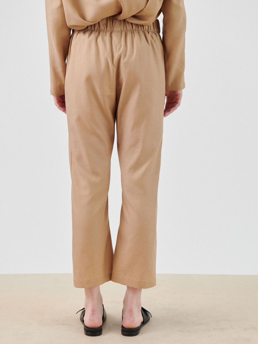 Back view of a person wearing beige pants and a matching long-sleeve top. The Eko Pant by Zero + Maria Cornejo is ankle-length with an elastic waistband. The person is standing on a beige surface, and only their lower half is visible. They are also wearing black slide sandals.