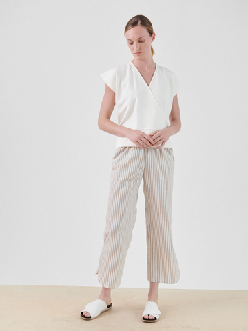 A person in a white V-neck top, Ruched Front Pant by Zero + Maria Cornejo, and white slide sandals stands on beige flooring against a plain white backdrop, looking down with hands gently clasped.