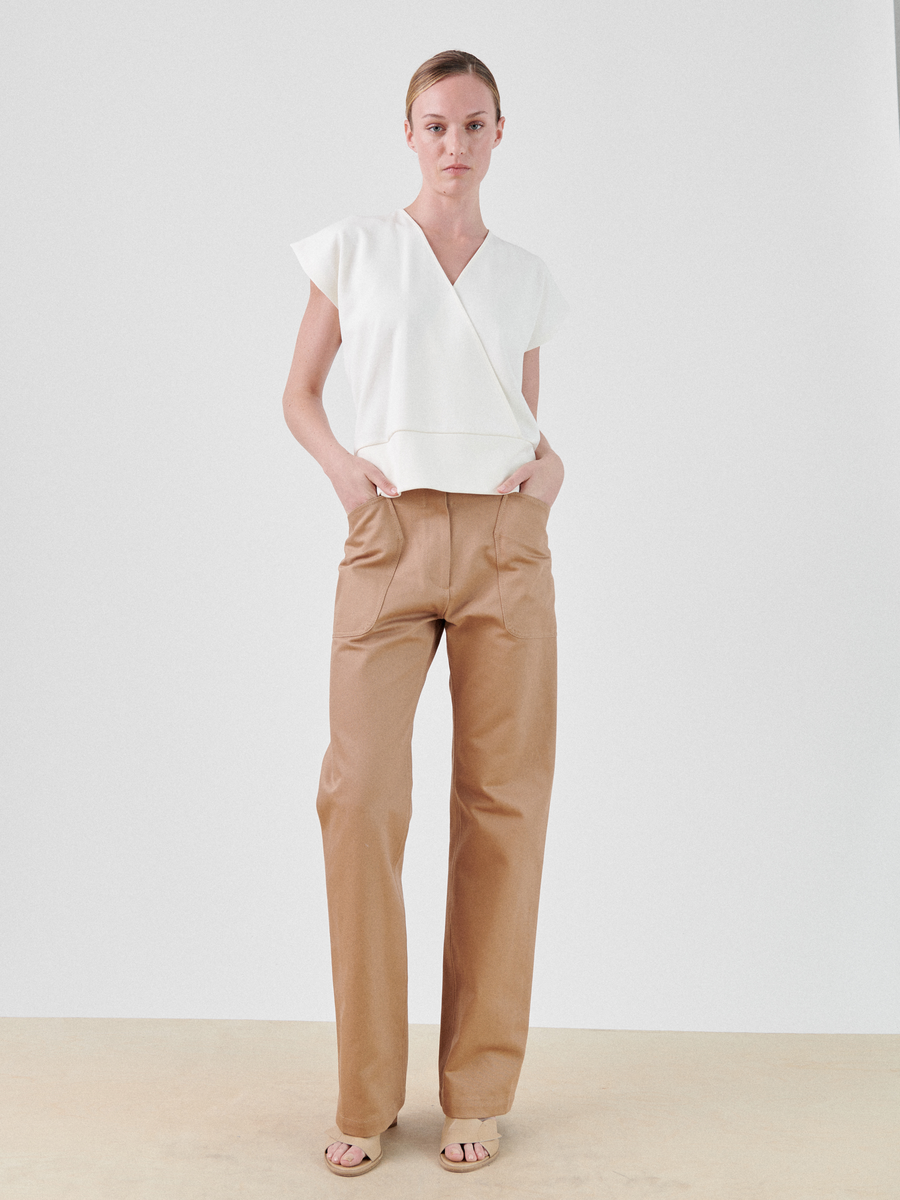 A person stands against a plain background, wearing a white, short-sleeve, Zero + Maria Cornejo Wrap Mido Top with an asymmetrical wrap design. They have on tan wide-leg high-waisted pants and beige open-toe sandals. Their hands are casually placed in their pockets.