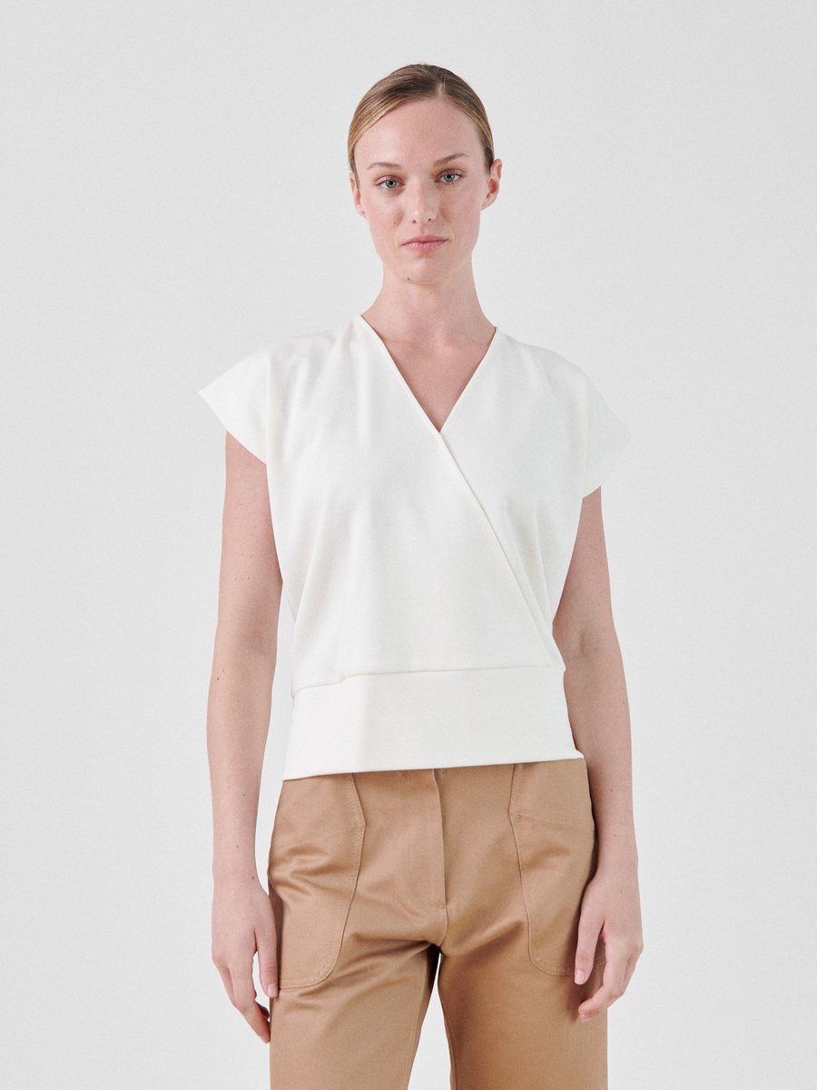 A person with short blonde hair is wearing a white Zero + Maria Cornejo Wrap Mido Top and light brown pants. They are standing against a plain white background and looking directly at the camera with a neutral expression.