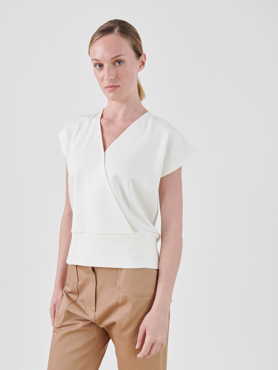 A woman with light skin and blond hair in a ponytail is wearing a white, short-sleeve, Wrap Mido Top by Zero + Maria Cornejo and tan trousers. She stands against a plain white background with her hands resting by her sides. She has a neutral expression on her face.