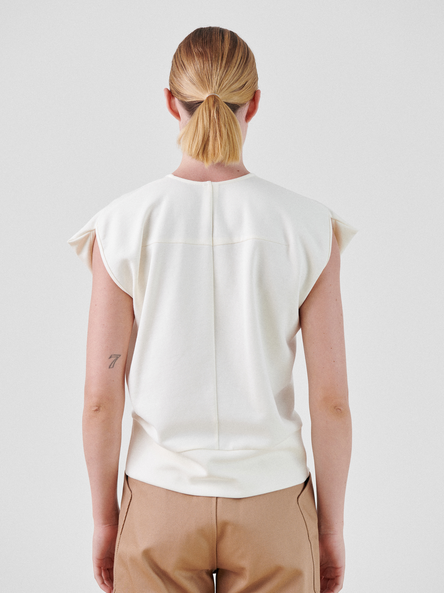A person with blonde hair tied in a low ponytail is shown from the back. They are wearing a sleeveless, white Wrap Mido Top by Zero + Maria Cornejo with folded cuffs, and brown pants. The person has a small tattoo on their left upper arm. The background is plain white.