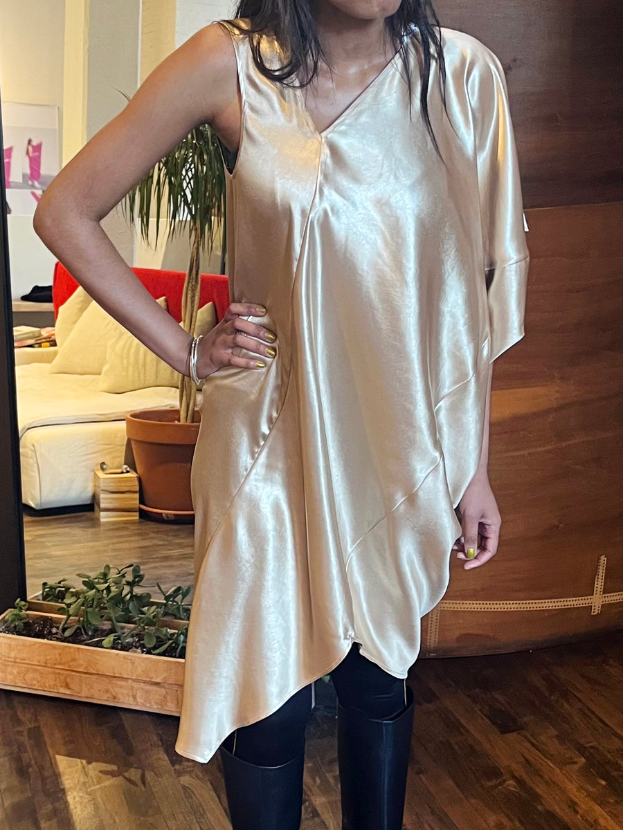 Wearing Zero + Maria Cornejo's Long Cade Dress, featuring a shiny metallic gold and asymmetrical design with one sleeve, the person pairs it with black knee-high boots. A living area with a couch and potted plant is visible in the background.